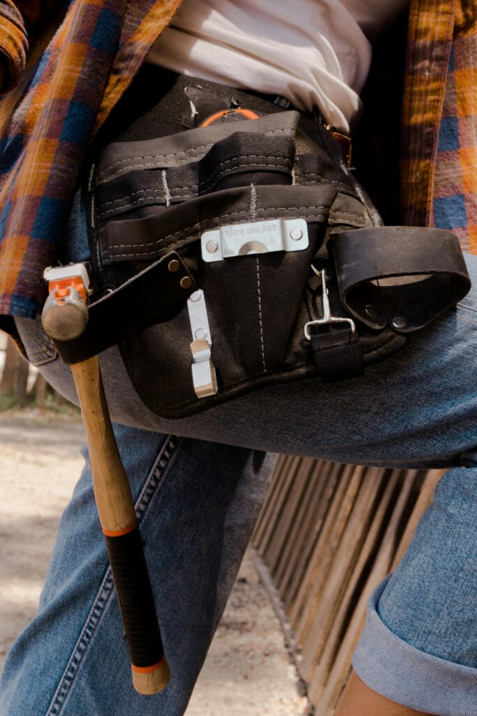 Close-up of a utility belt filled with tools on a person wearing jeans and a flannel shirt outdoors.